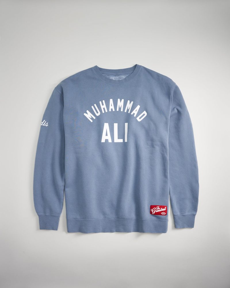 A light blue sweatshirt featuring the name Muhammad Ali prominently displayed in bold white letters across the front with a red tag at the bottom reading The Greatest