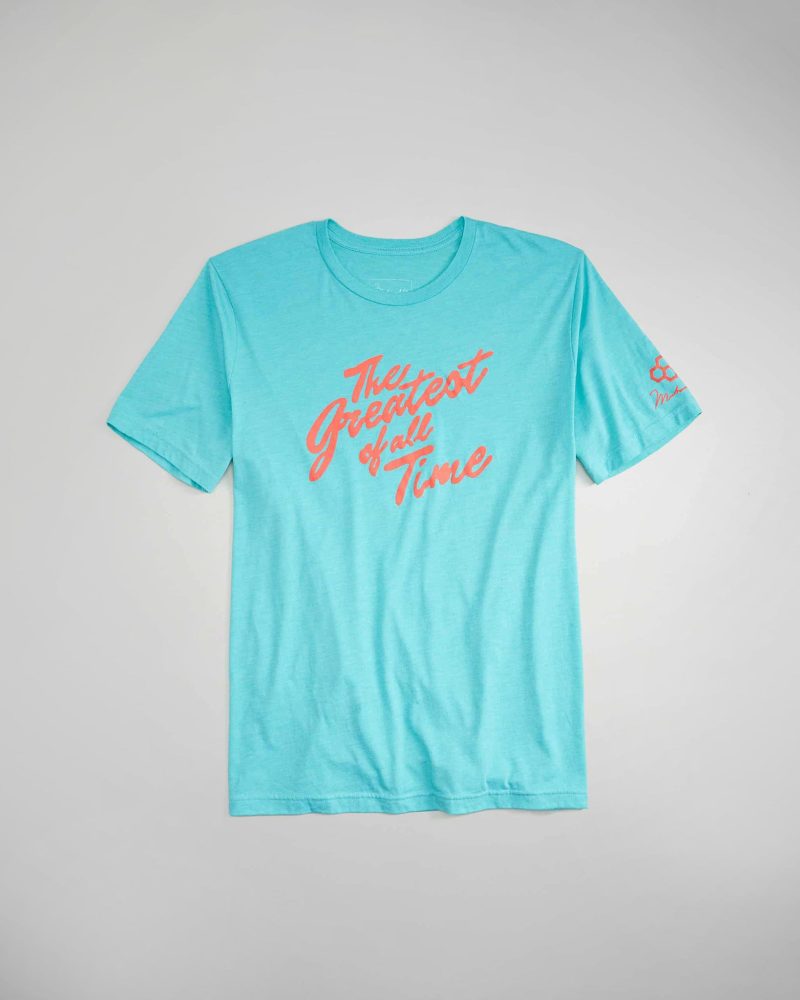 A light blue t-shirt featuring the phrase The Greatest of All Time in bold coral lettering designed for casual wear