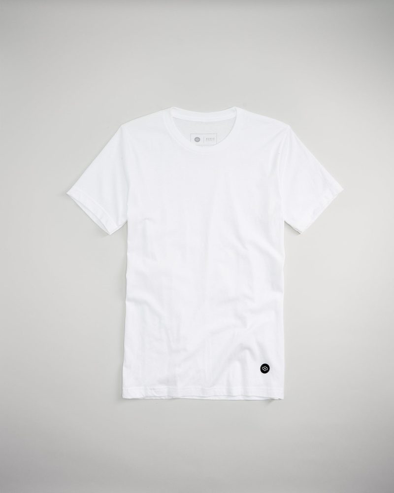 A plain white t-shirt displayed flat against a muted gray background featuring a subtle logo on the bottom left