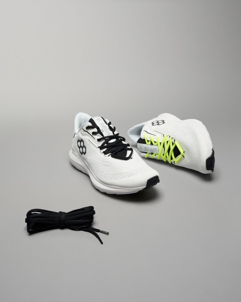 A pair of modern running shoes in white with contrasting black and neon yellow laces set against a neutral background featuring textured mesh uppers and a sleek design