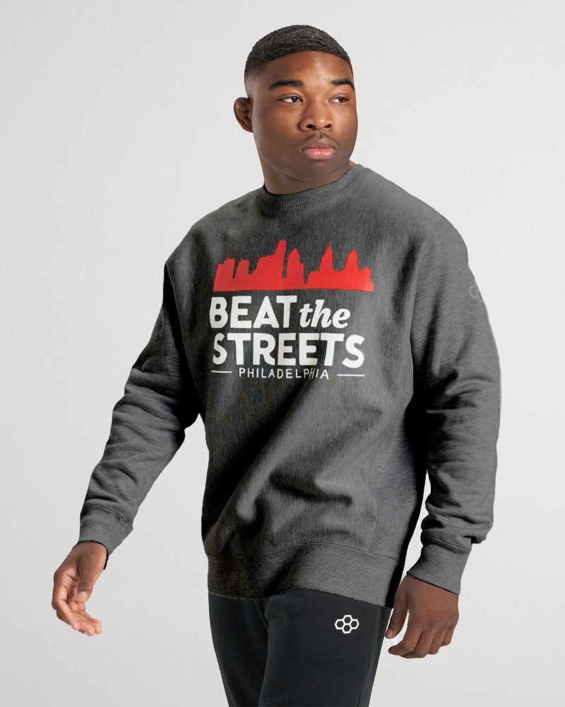 A young man wearing a gray sweatshirt with the phrase BEAT the STREETS PHILADELPHIA and black pants featuring a white logo posing confidently against a soft gray background